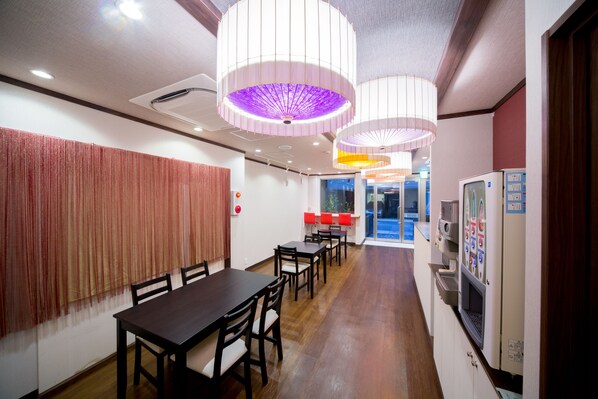 Un comedor con una mesa larga y sillas, una máquina expendedora y vistas al exterior.