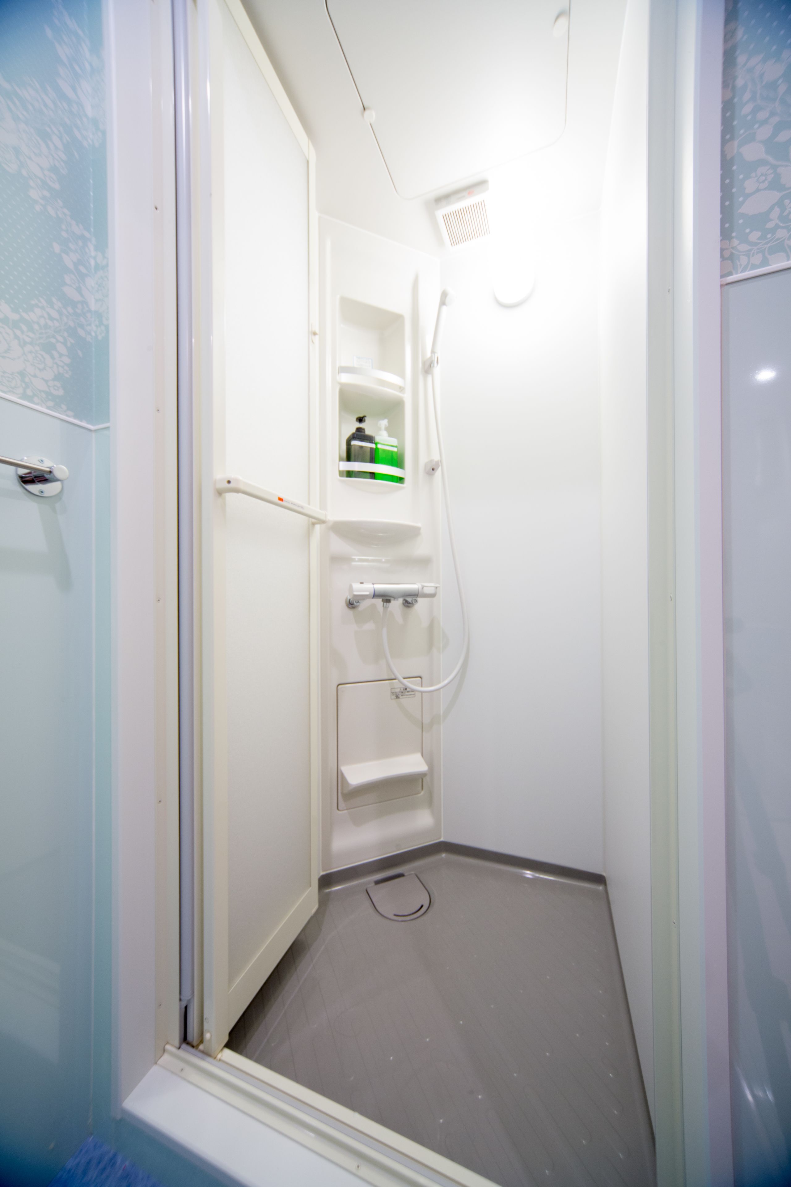 A small, enclosed shower stall with a white door, a shelf with toiletries, and a showerhead.