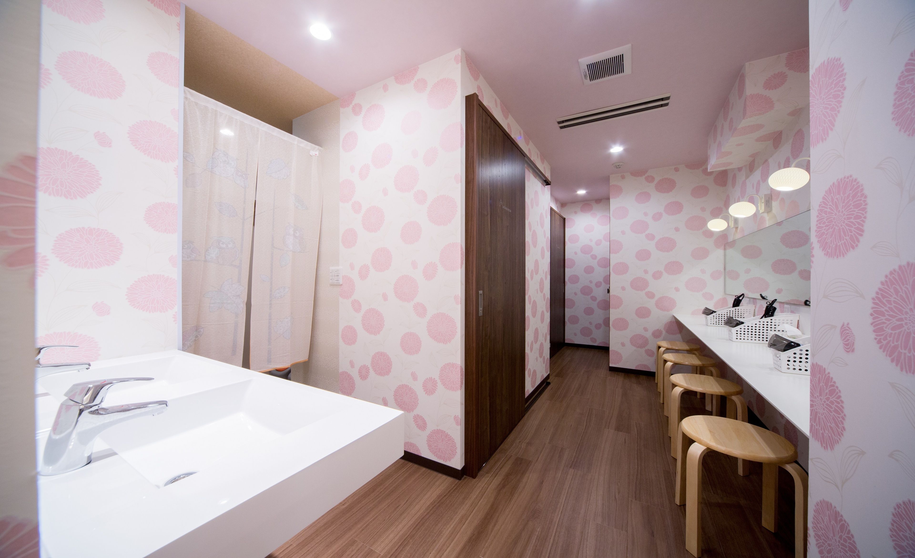 Baño con un lavamanos grande, taburetes de madera, un espejo y papel tapiz con estampado floral rosa.