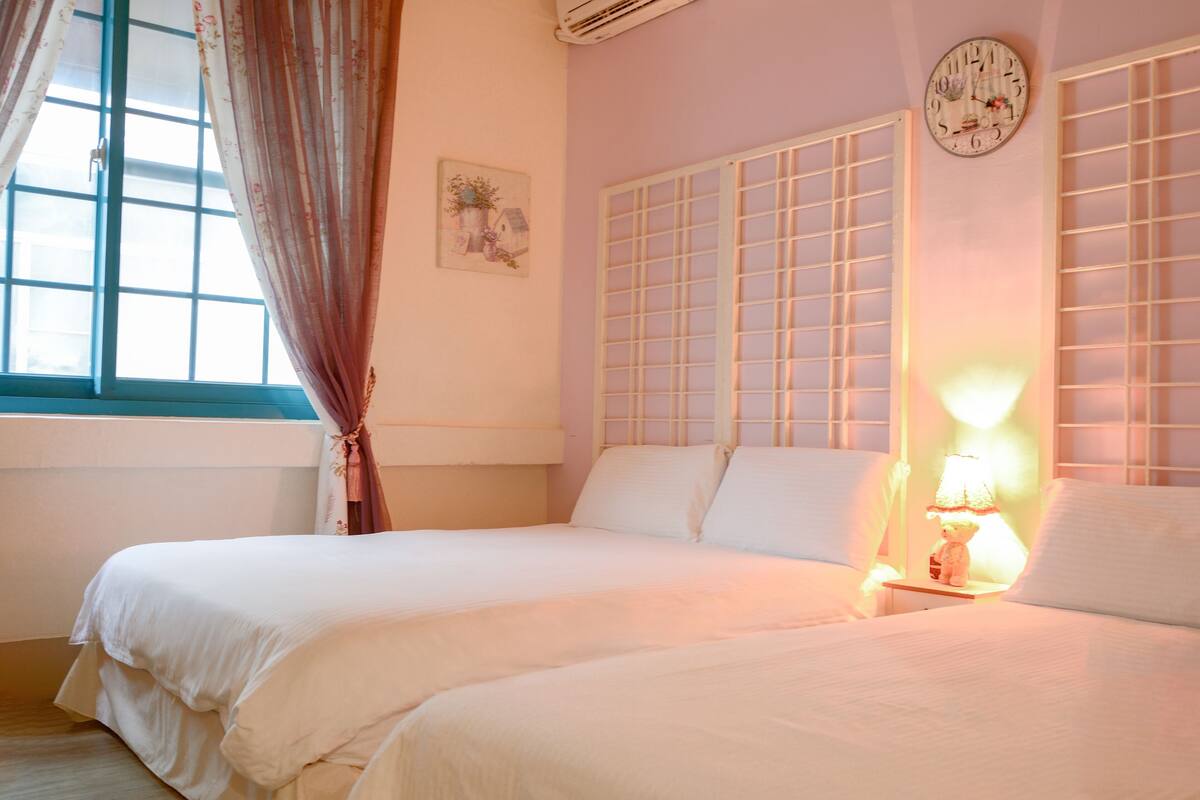 A bedroom with two beds, a window with curtains, a wall clock, and a bedside lamp.