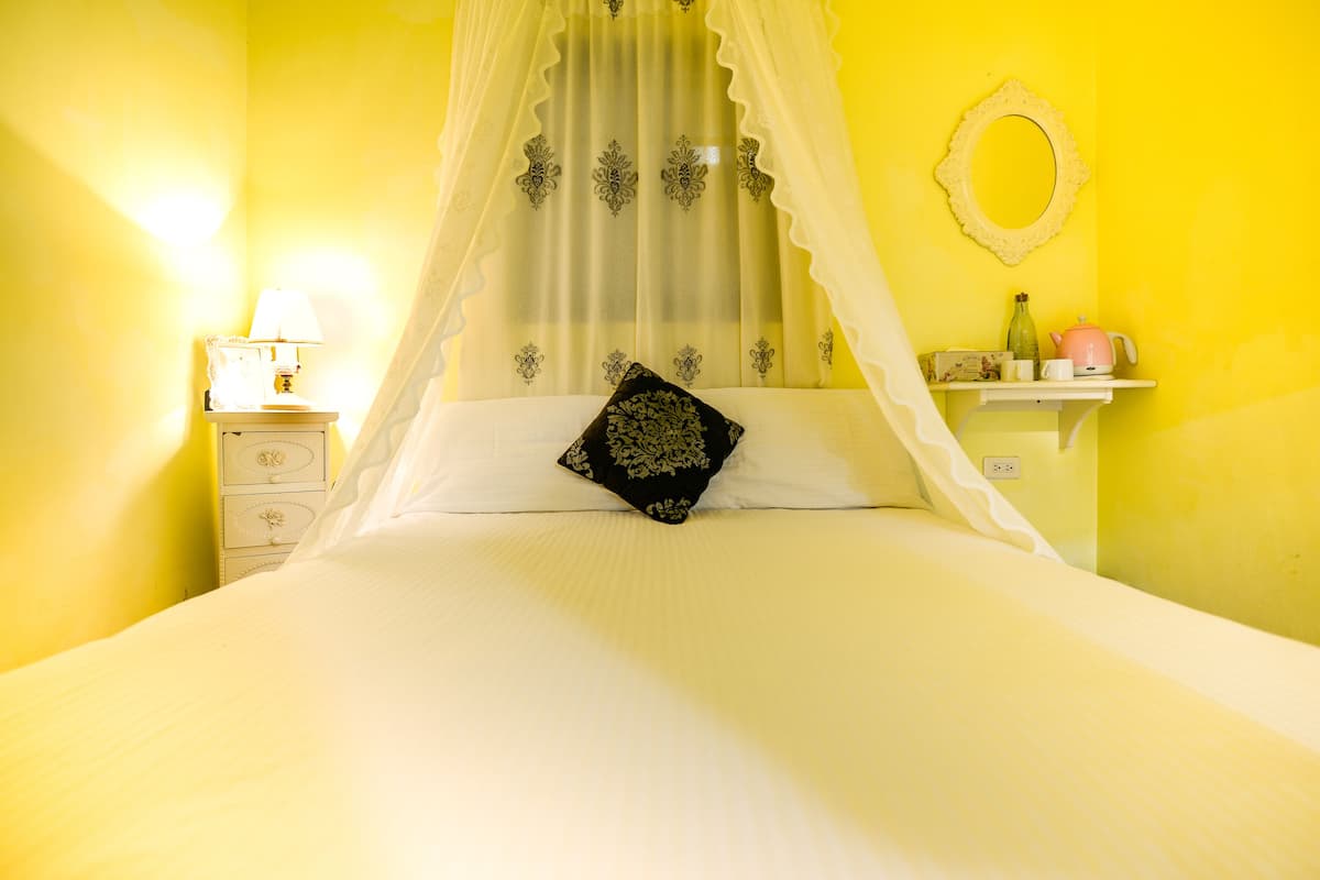 A bed with a canopy, a bedside table with a lamp, and a small shelf with a mirror and a pink kettle.