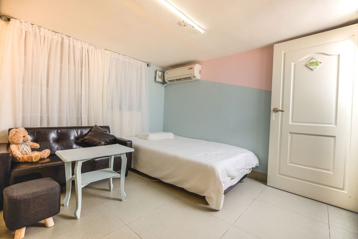 A room with a bed, a small table, a chair, a teddy bear, and an air conditioning unit.