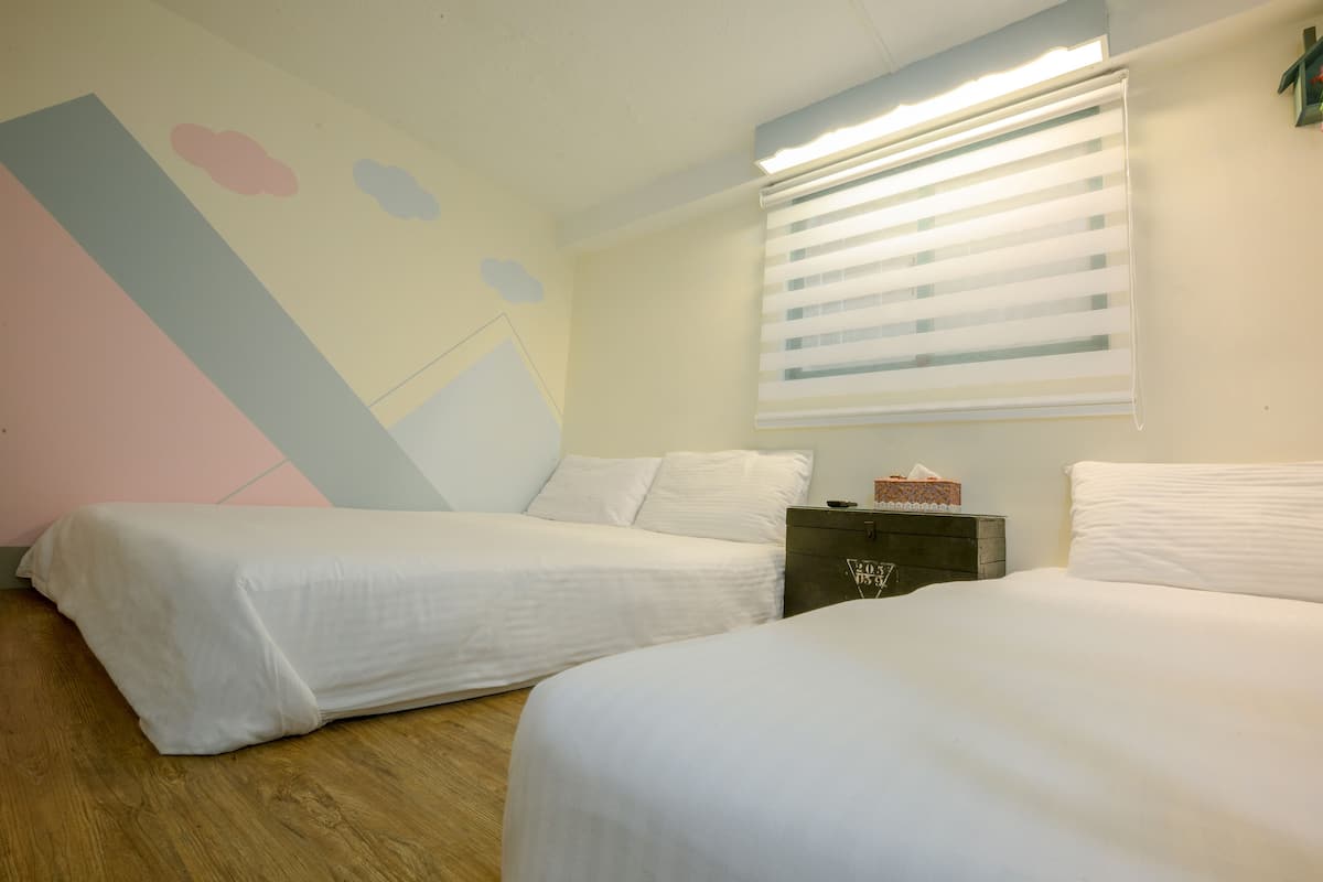 A bedroom with two single beds, a wooden floor, a wall with a geometric design, a window with blinds, and a bedside table with a tissue box.