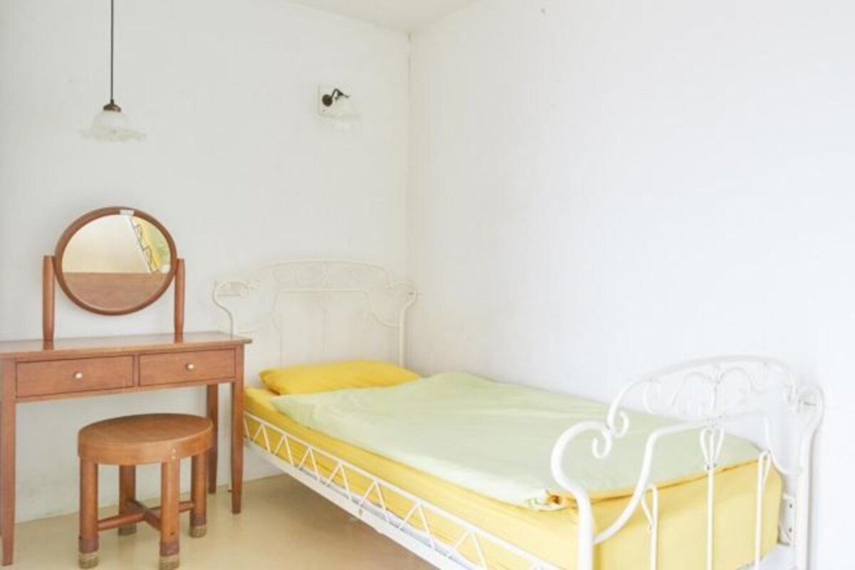 A small, single-bed room with a wooden desk and chair, a round mirror, and a white metal bed frame.