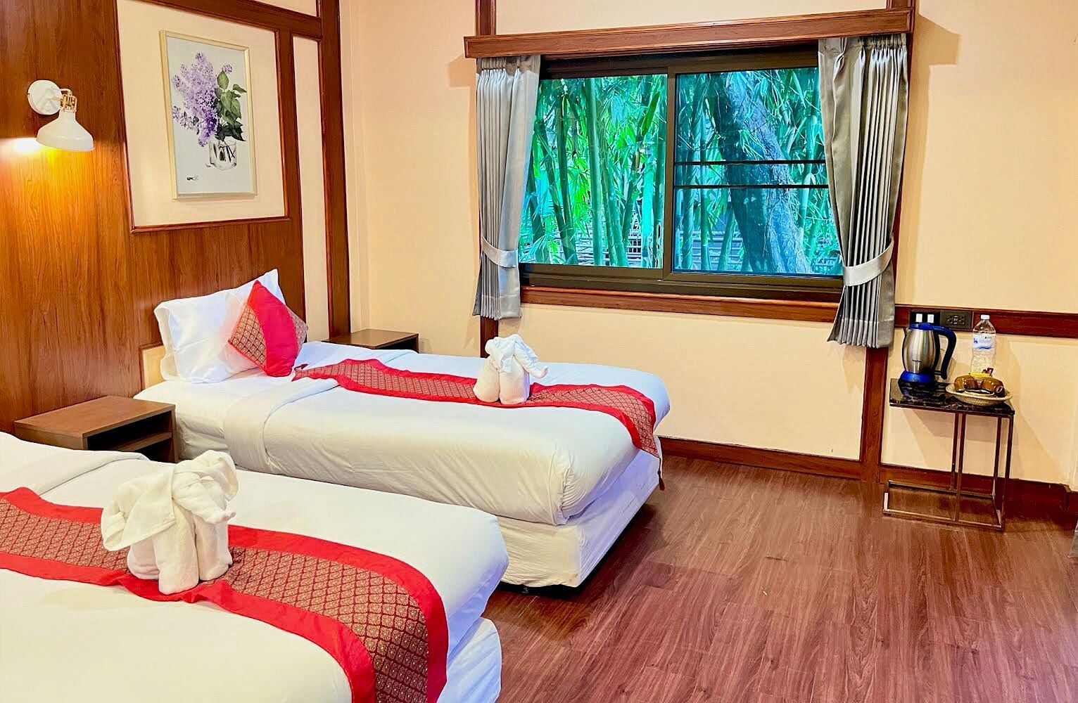 Una habitación de hotel con dos camas, una mesita de noche, una ventana con cortinas y una pequeña mesa con hervidor y botella de agua.