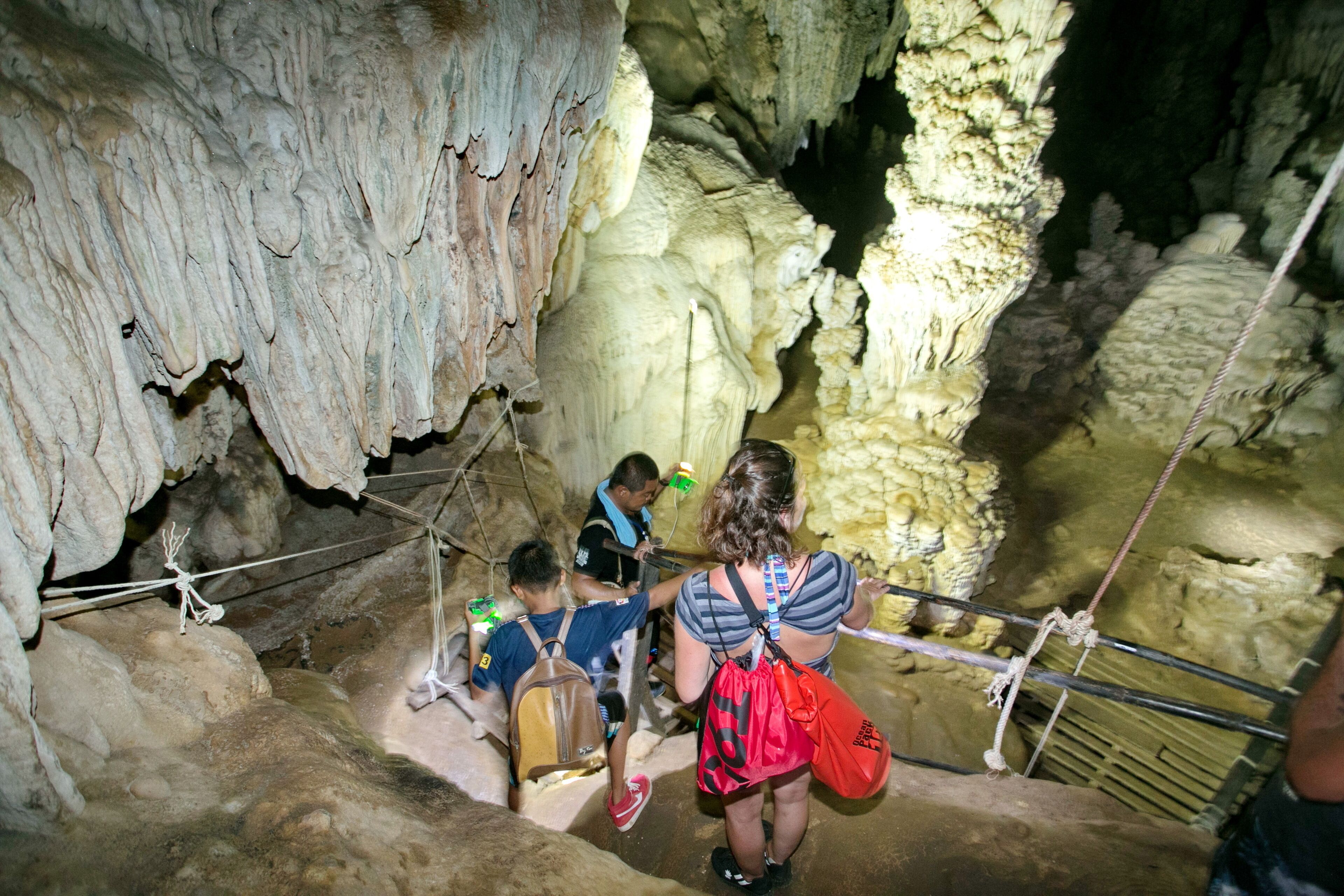 A cave with stalactites and stalagmites, a rope bridge, and visitors wearing backpacks.