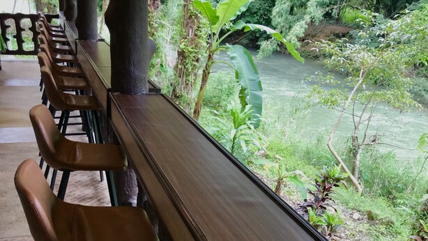 A row of wooden chairs and a bench facing a river with lush greenery.