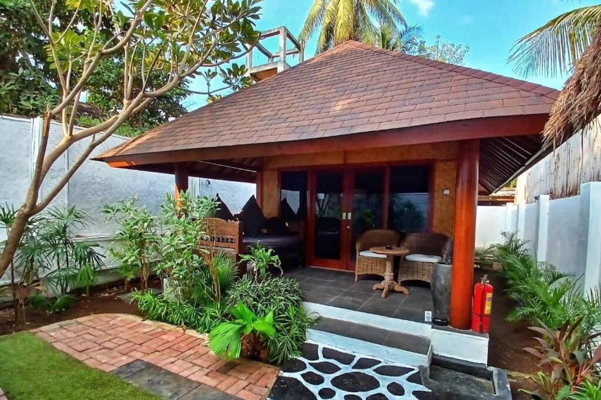 Een huis met een pannendak, een veranda met rieten meubels, en een stenen pad dat naar de ingang leidt.