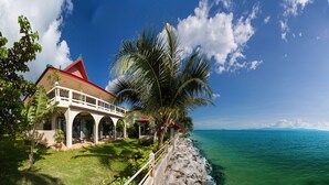 Une propriété située en bord de mer, avec un bâtiment blanc surmonté d’un toit rouge, un palmier et une vue sur l’océan.