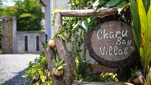 Une pancarte en bois portant l’inscription « Charu Bay Villas », entourée de verdure et d’une clôture rustique.