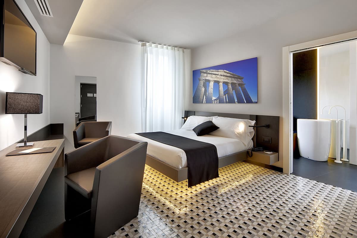 A modern hotel room with a bed, desk, chairs, a large window with curtains, a painting of a historical building, and a patterned floor.