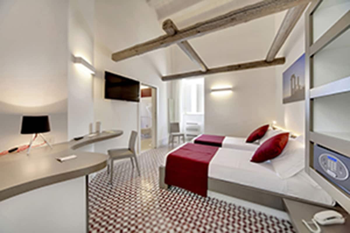 A modern hotel room with a bed, bedside table, chair, and a desk with a lamp.