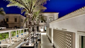 A modern outdoor lounge area with seating and palm trees, a covered walkway, and a building in the background. - Zafran Boutique Hotel (Scicli)