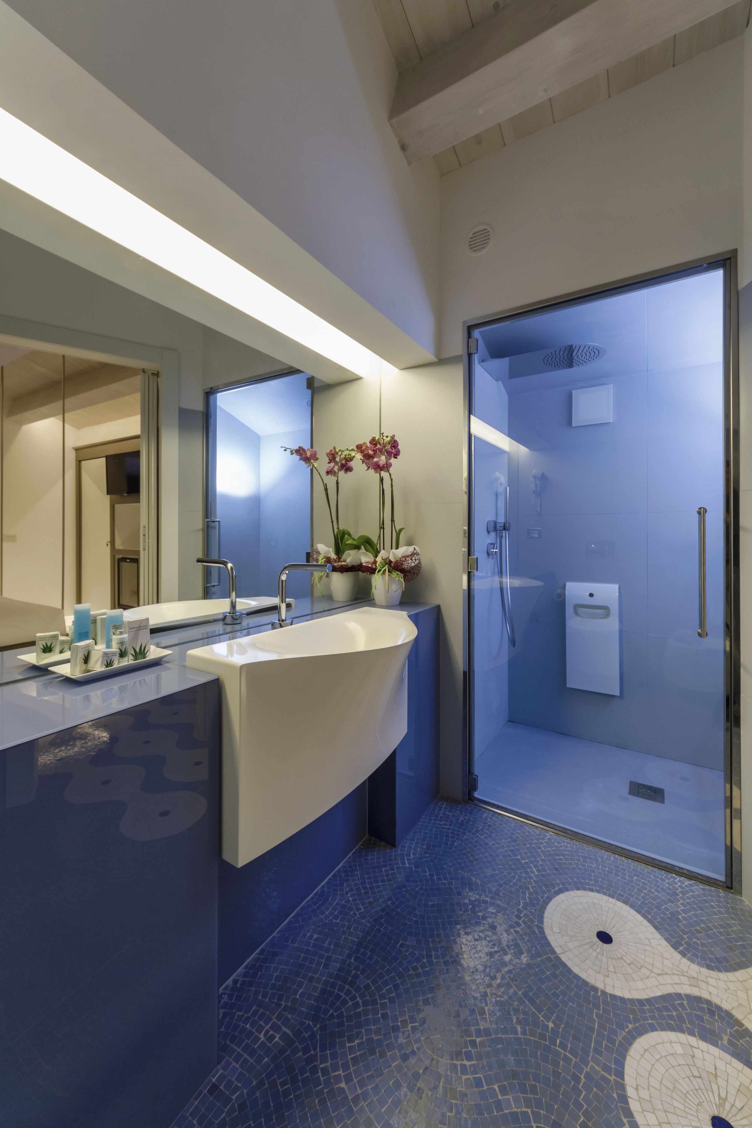 A modern bathroom with a large mirror, a white sink, a glass shower enclosure, and a blue tiled floor.