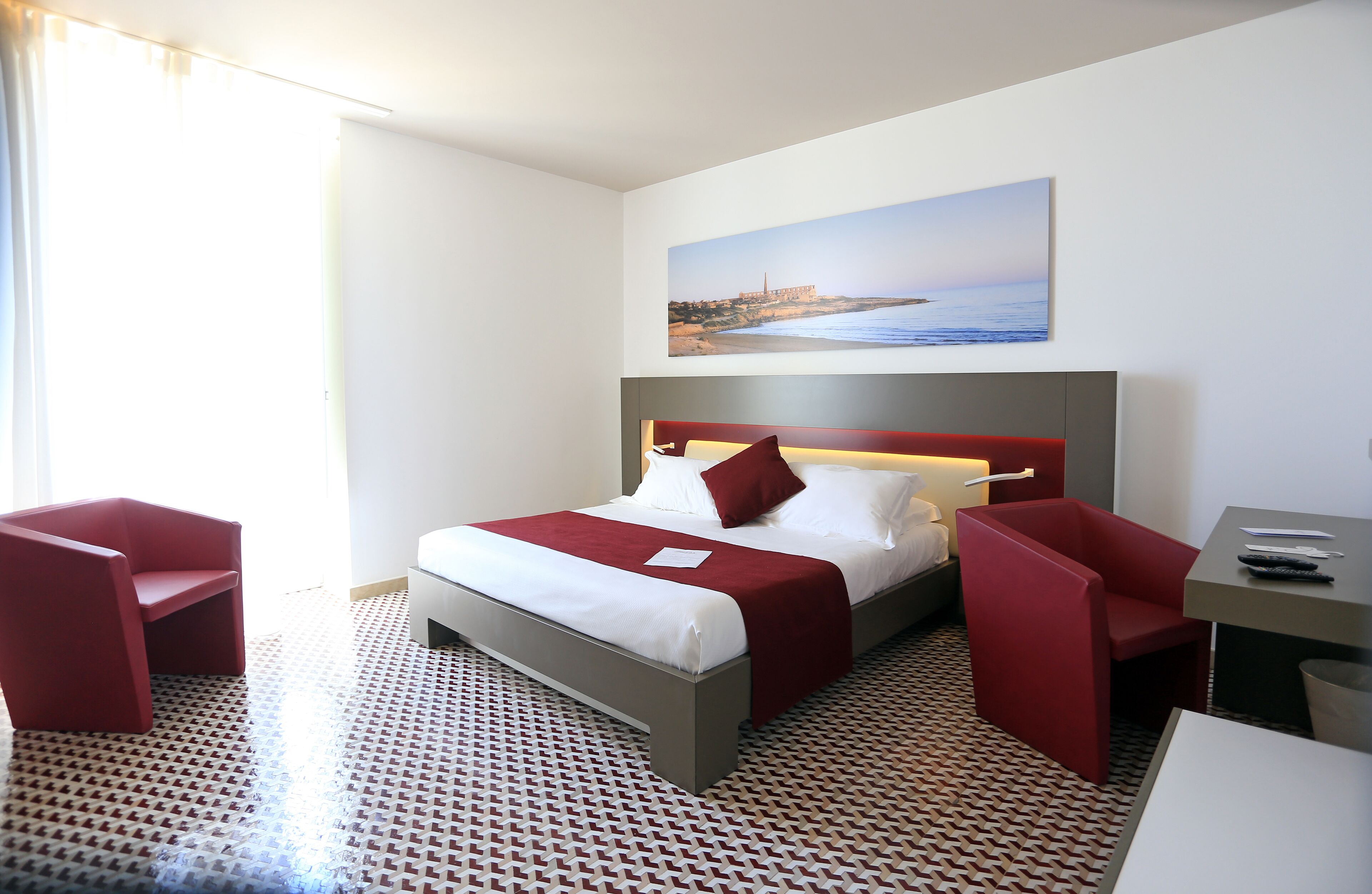 Ein Hotelzimmer mit einem Bett, zwei roten Sesseln, einem Schreibtisch und einem Gemälde einer Küstenlandschaft.