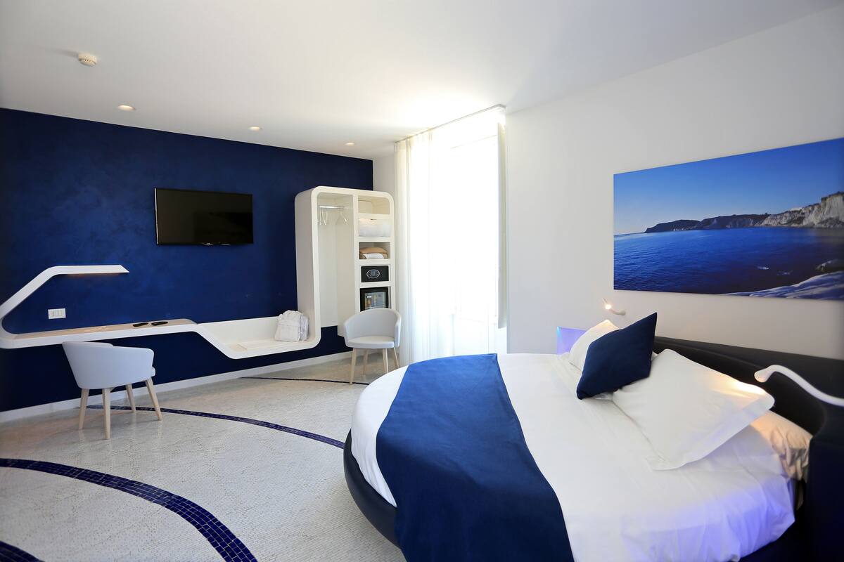 A modern hotel room with a large bed, a desk, a chair, and a wardrobe.
