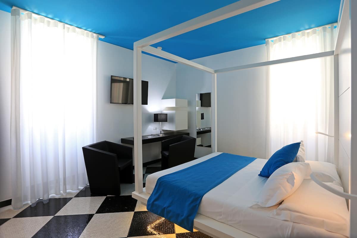 A hotel room with a four-poster bed, a desk with a chair, a mirror, and a window with sheer curtains.