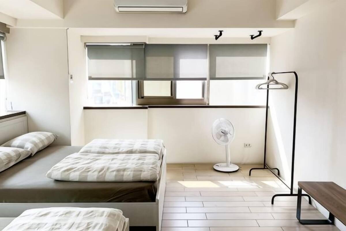 A bedroom with a bed, a bedside table, a fan, a bench, and a window with blinds.