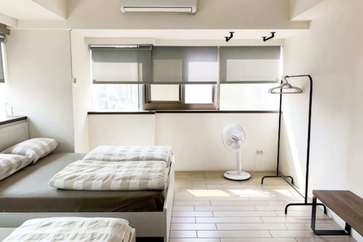 A bedroom with a bed, a bedside table, a fan, a bench, and a window with blinds.