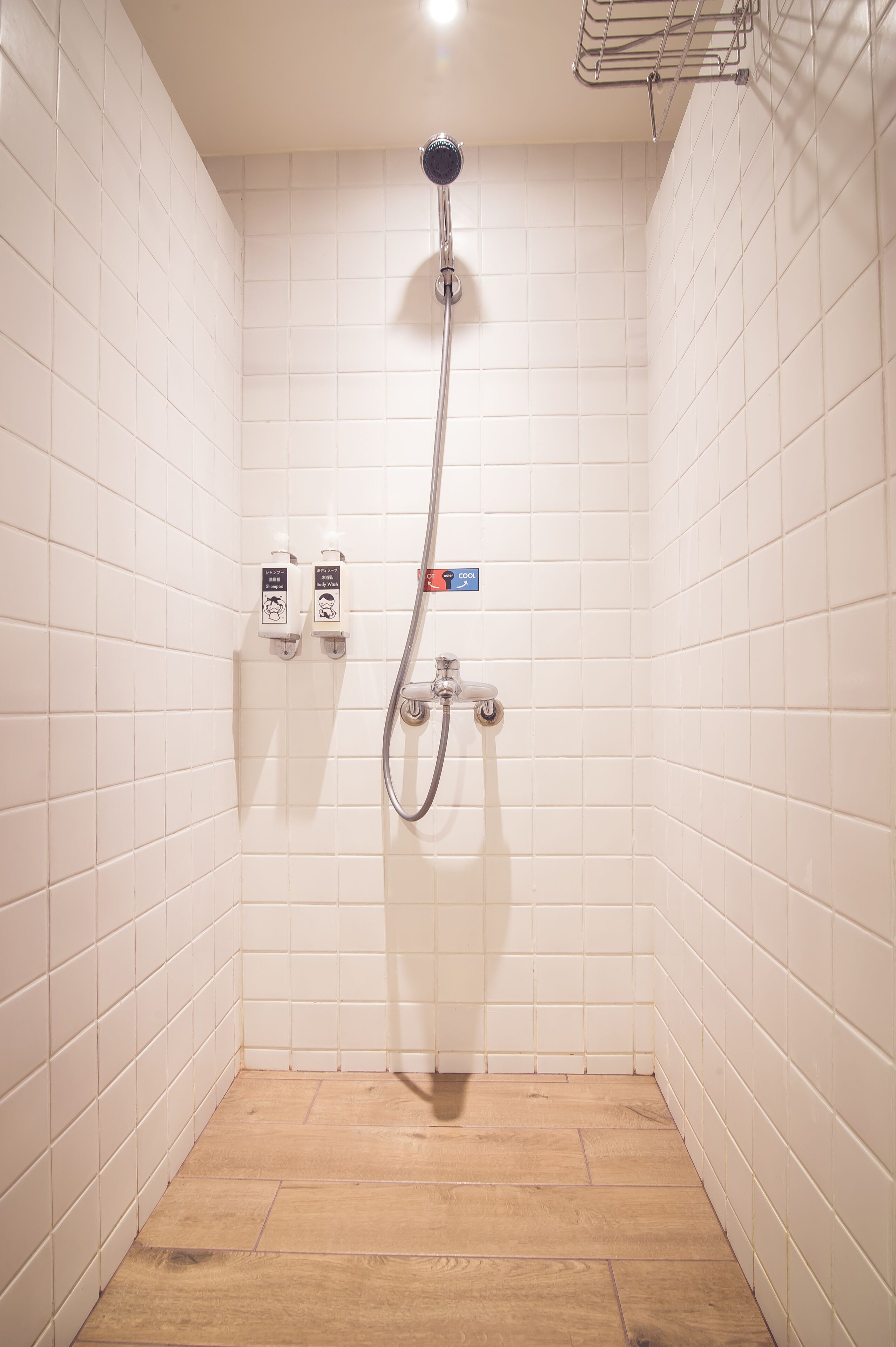 A small, tiled shower room with a wooden floor, a handheld showerhead, and two wall-mounted soap dispensers.
