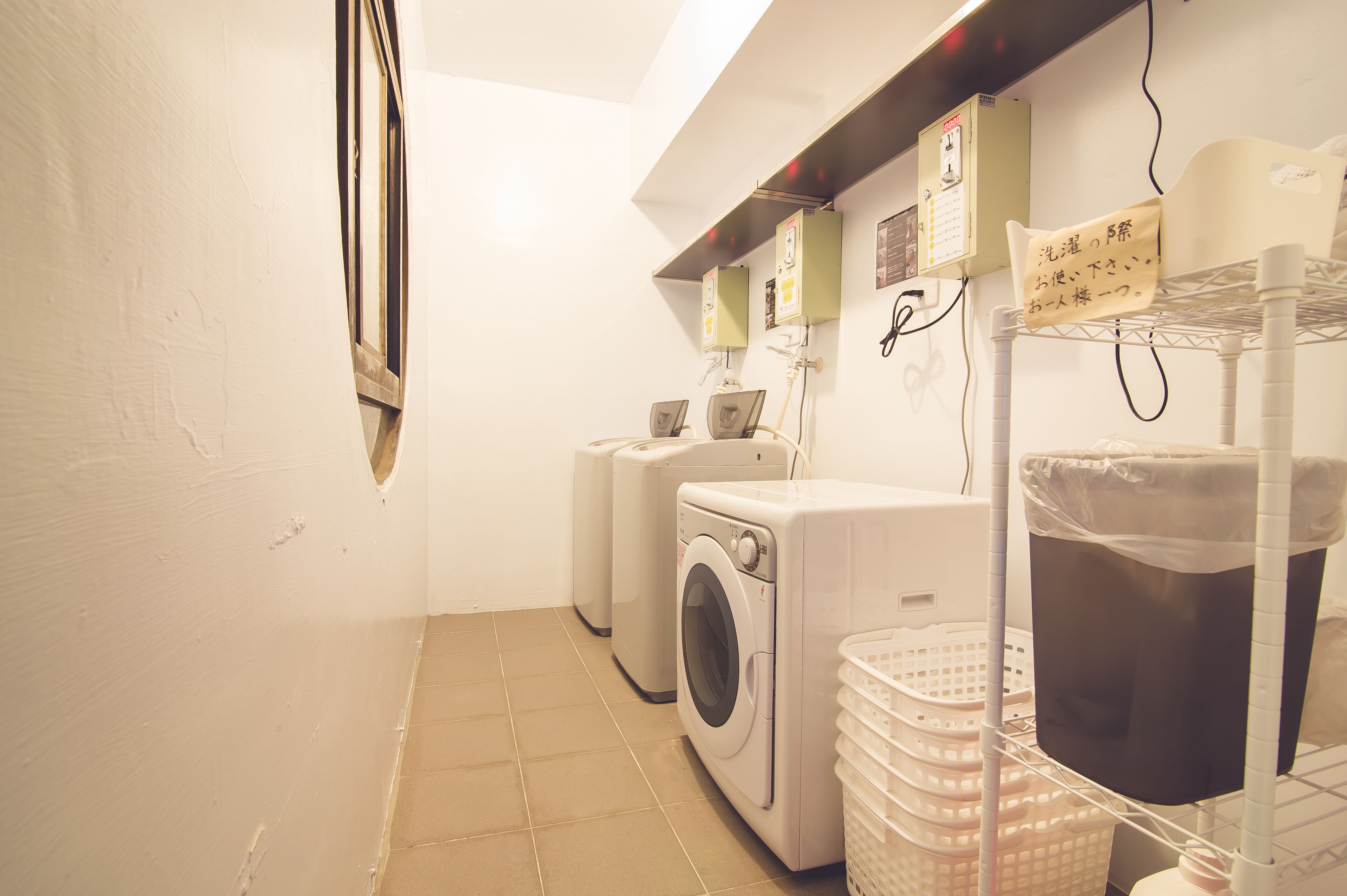 A laundry room with two washing machines, a drying machine, and a shelf with baskets and a trash bin.