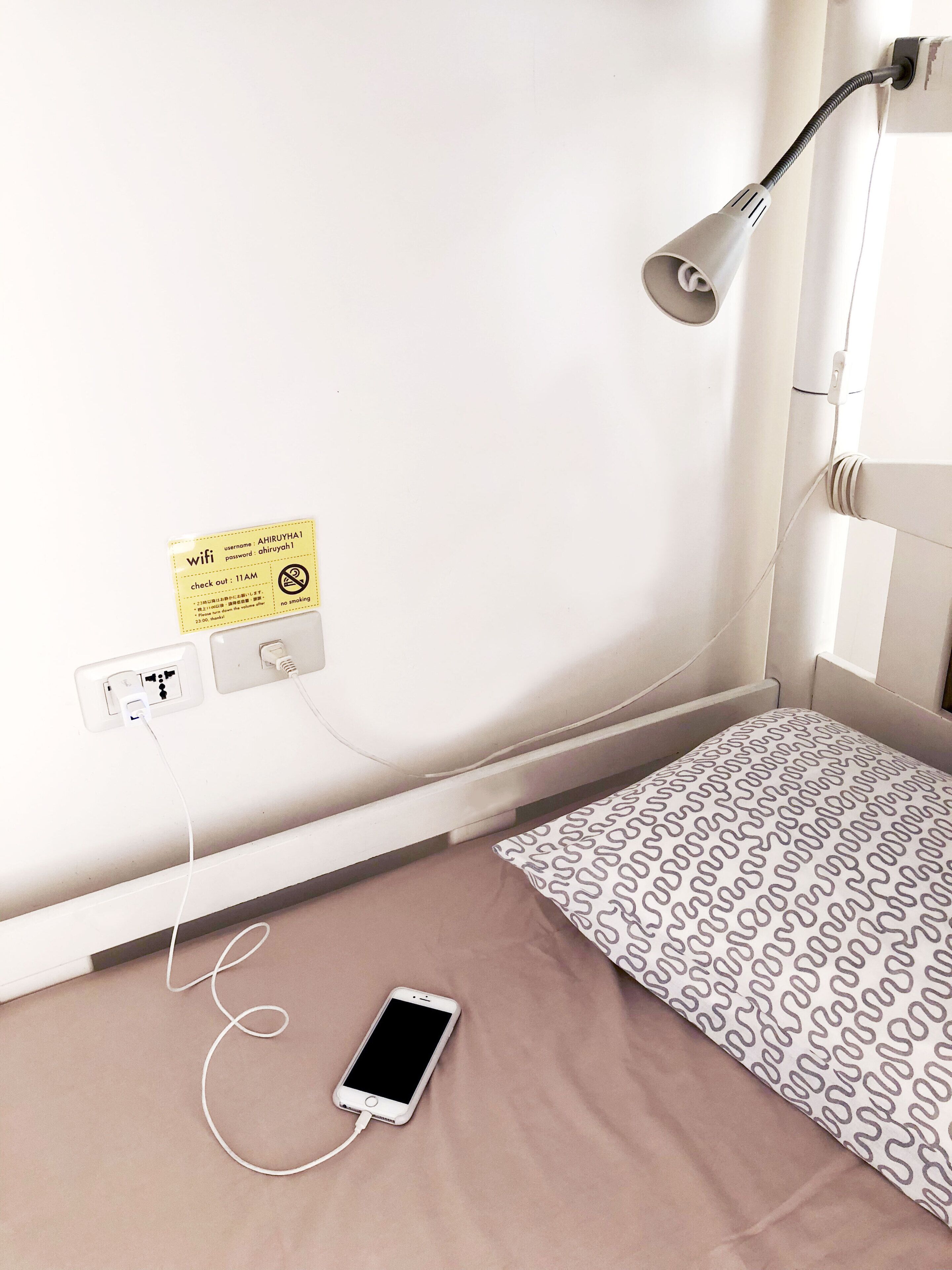 Quarto de hotel com cama, luminária de mesa, smartphone carregando, tomada e sinal de senha wifi.