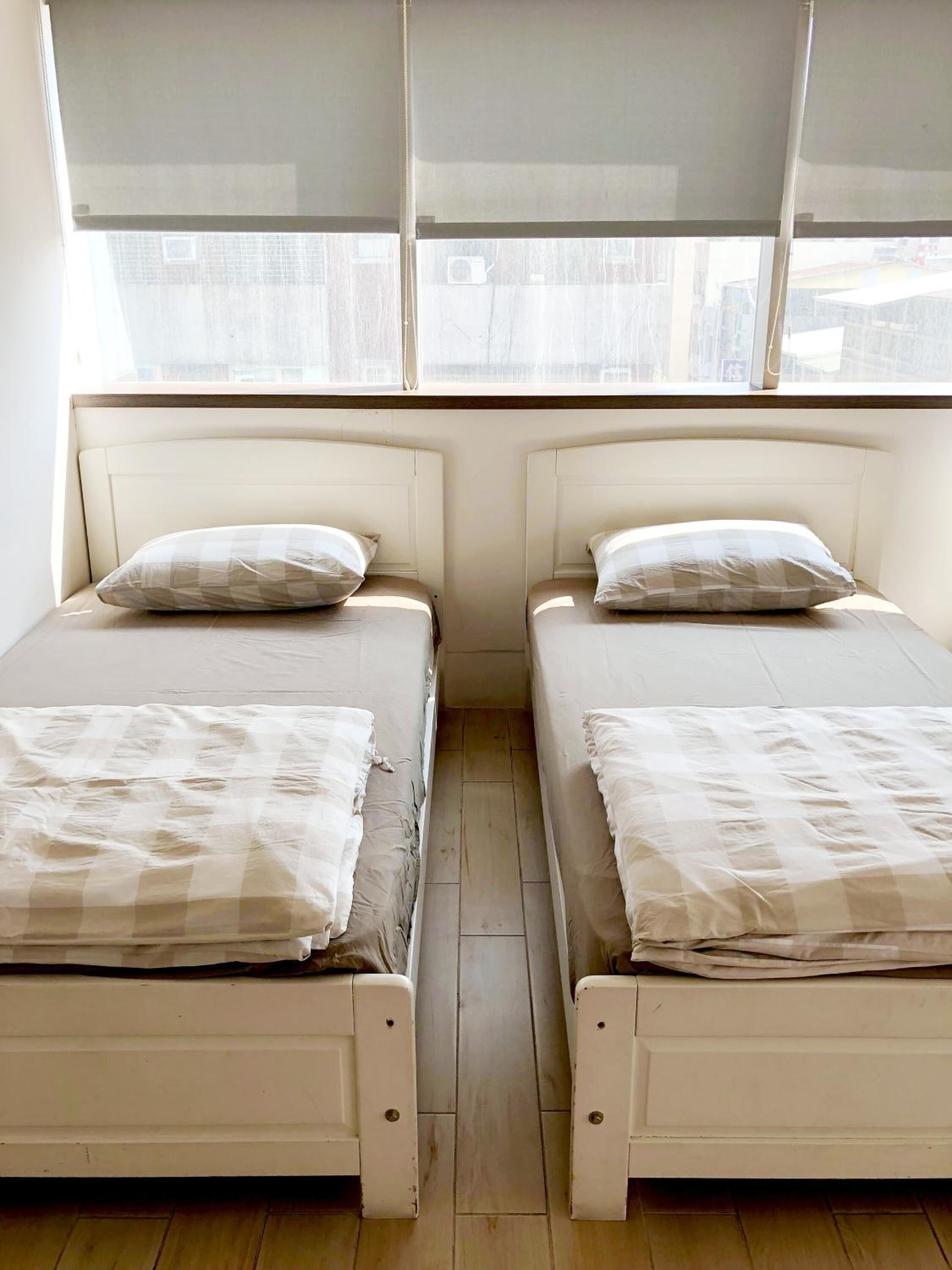 A room with two single beds, each with a pillow and a blanket. The beds are positioned against a wall with a window above them, allowing natural light to enter the room.