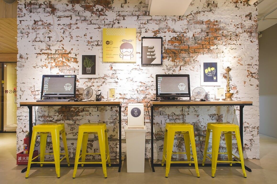 Ein Arbeitsplatz mit zwei Schreibtischen, jeweils mit einem Laptop und einem Ventilator, sowie drei gelben Hocker.