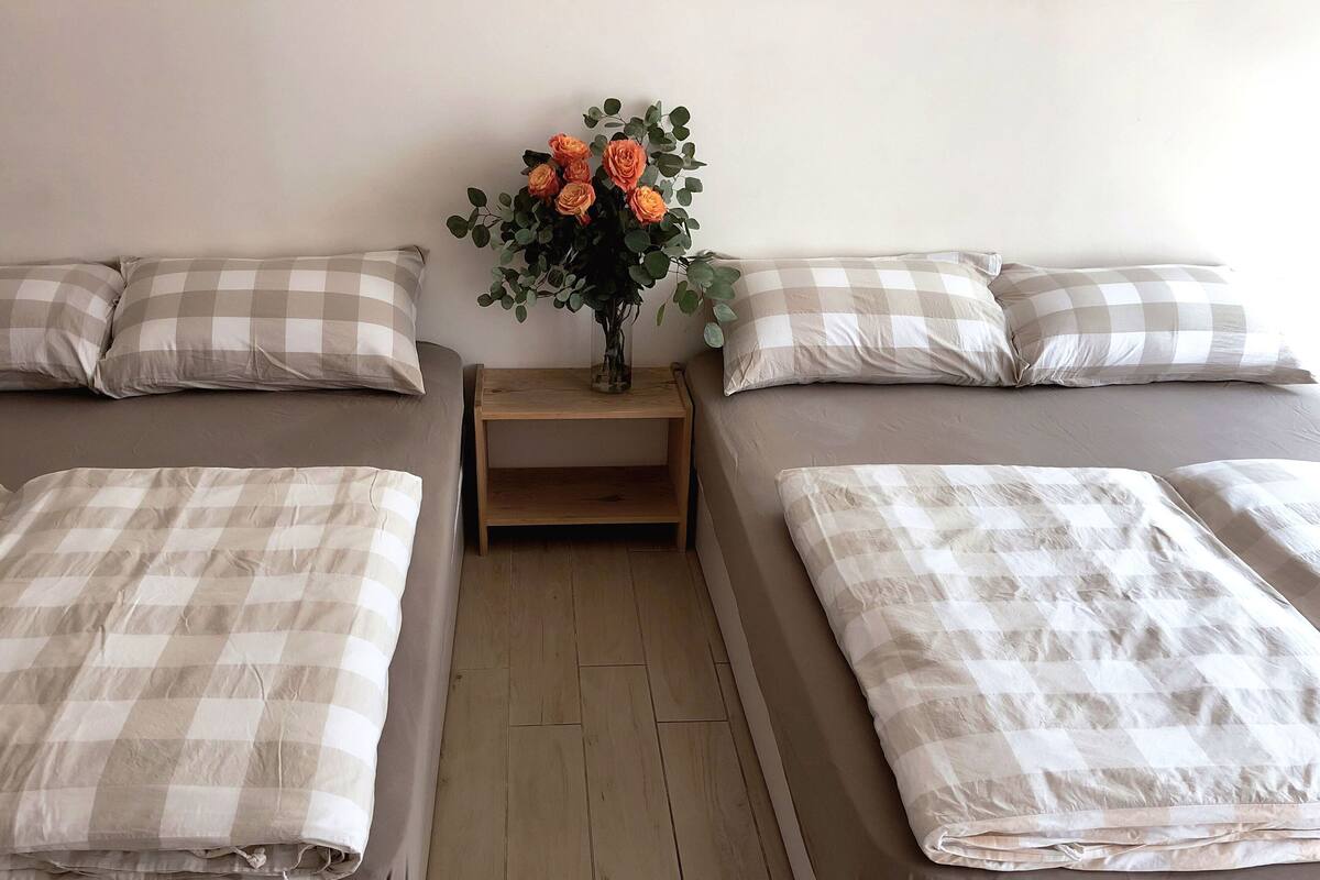 A hotel room with two beds, each with a checkered bedspread and matching pillows. There is a wooden nightstand between the beds with a vase of flowers on it.