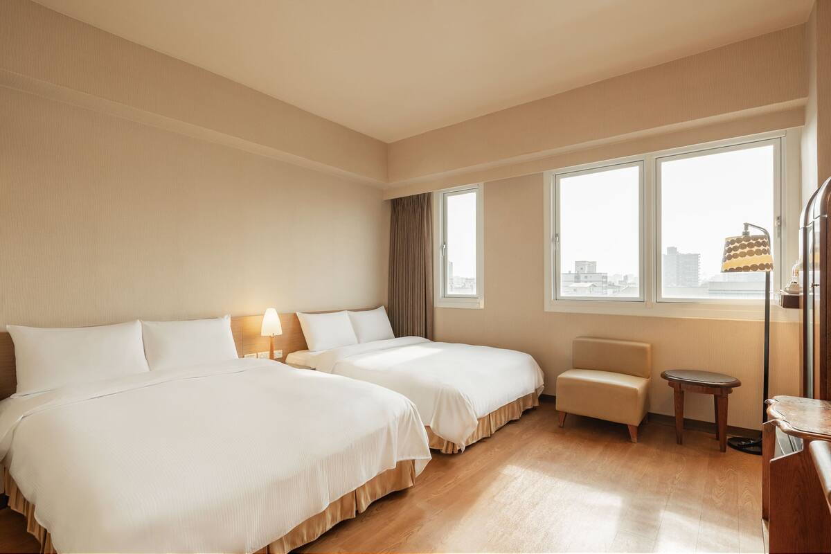 Habitación de hotel con dos camas, una zona de estar pequeña y vistas a la ciudad a través de amplias ventanas.