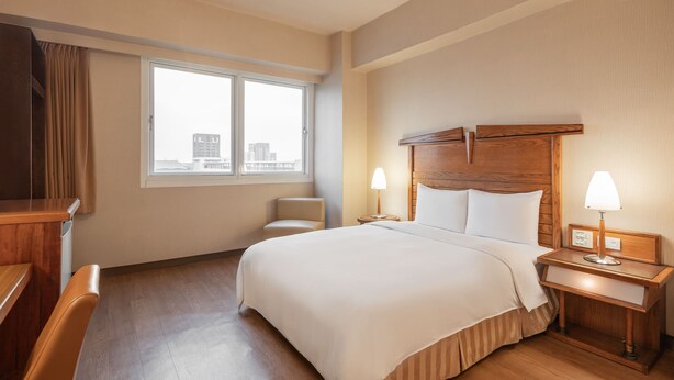 Habitació d'hotel amb un llit gran, dues làmpades de nit, una tauleta de nit de fusta, una cadira i una finestra amb vista a edificis.