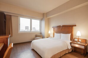 Une chambre d’hôtel avec un grand lit, deux lampes de chevet, une table de nuit en bois, une chaise et une fenêtre donnant sur des immeubles.
