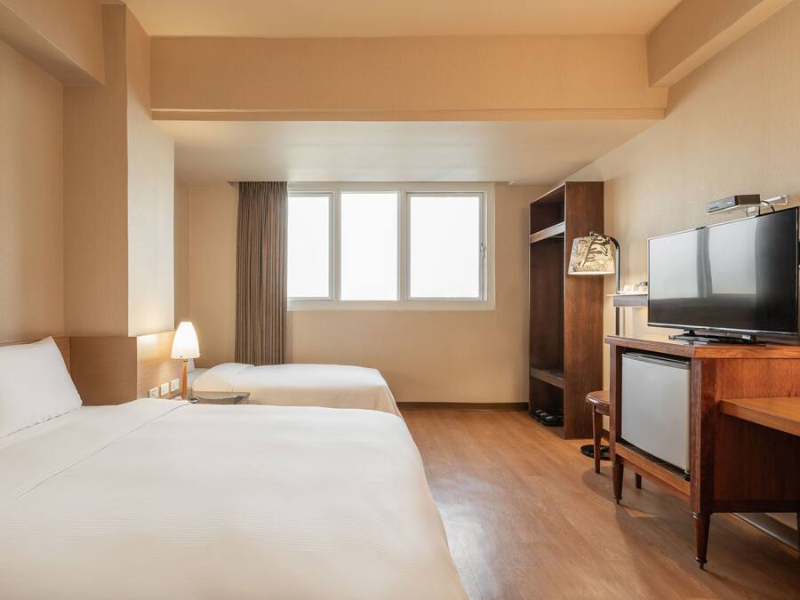 Ein Hotelzimmer mit zwei Betten, einem Fernseher, einem Schreibtisch und einem fensterbepflanzten Fenster.