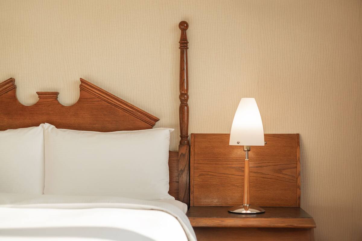 Una cama bien hecha con sábanas y almohadas blancas, una mesita de noche de madera con una lámpara y un cabecero de madera.