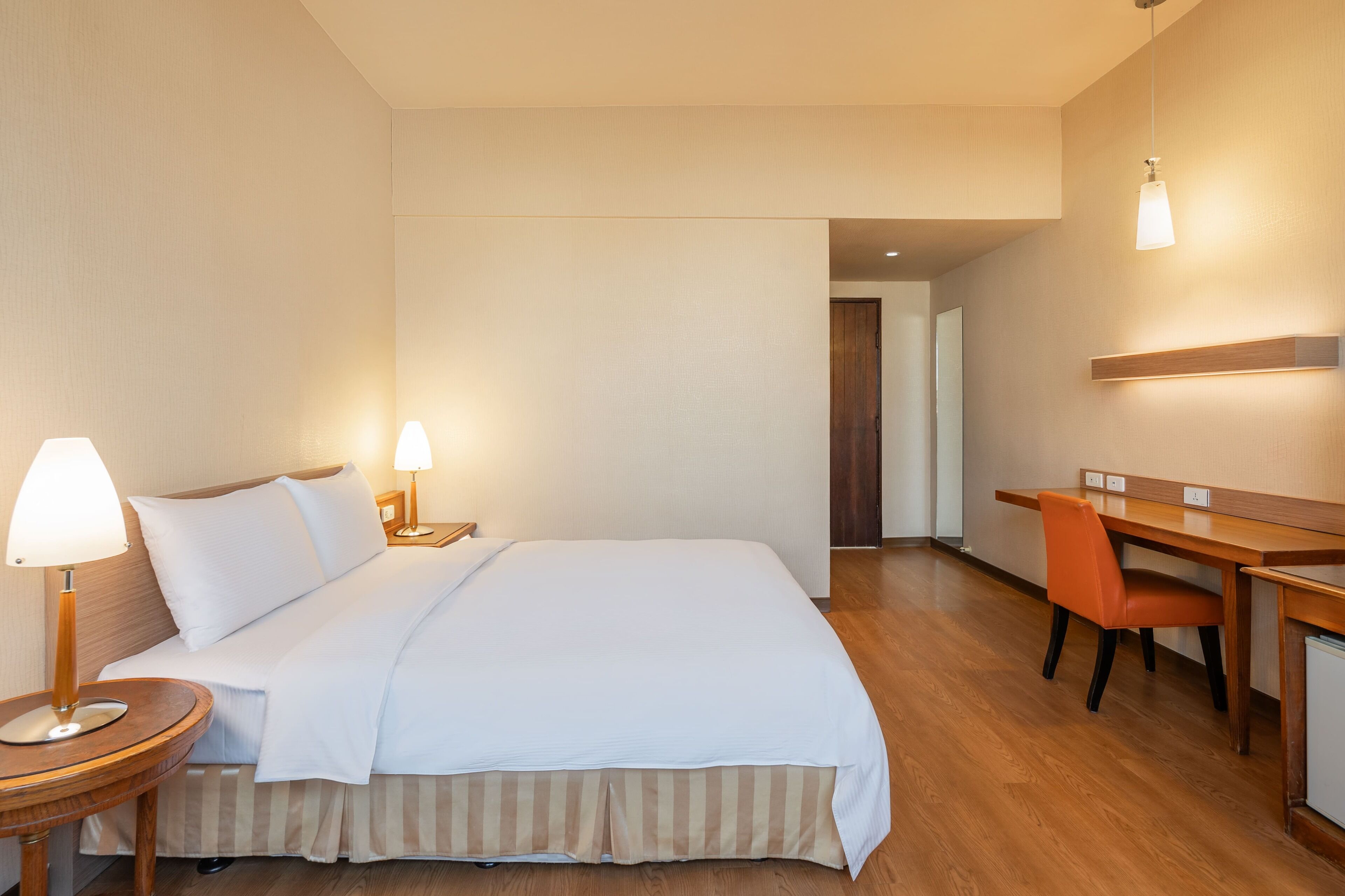 A hotel room with a bed, bedside lamps, a desk, and an orange chair.