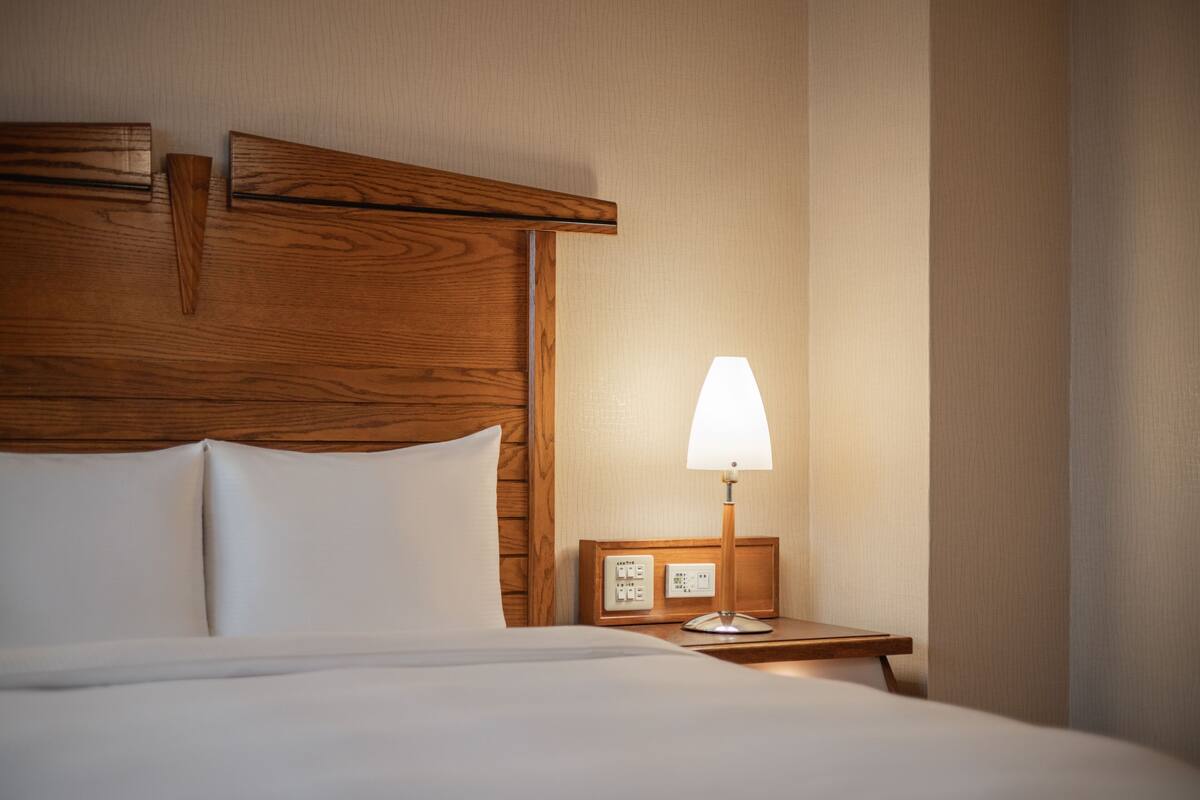 Habitación de hotel con cabecera de madera, cama con ropa blanca, mesita de noche con lámpara y panel de control.