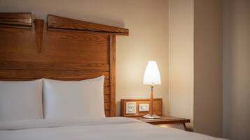 Habitación de hotel con cabecera de madera, cama con ropa blanca, mesita de noche con lámpara y panel de control.