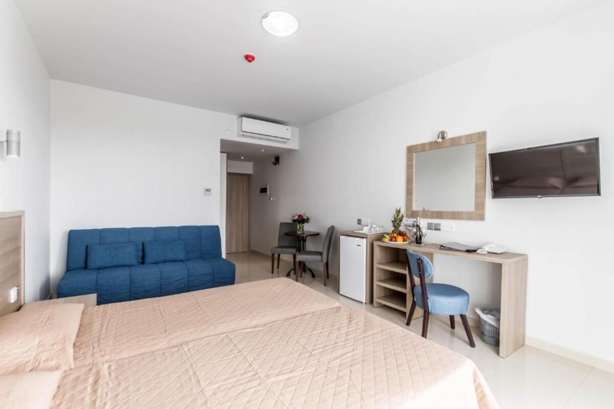 Ein Hotelzimmer mit einem großen Bett, einem Schreibtisch mit Stuhl, einer kleinen Kühlschrank und einem an der Wand befestigten Fernseher.