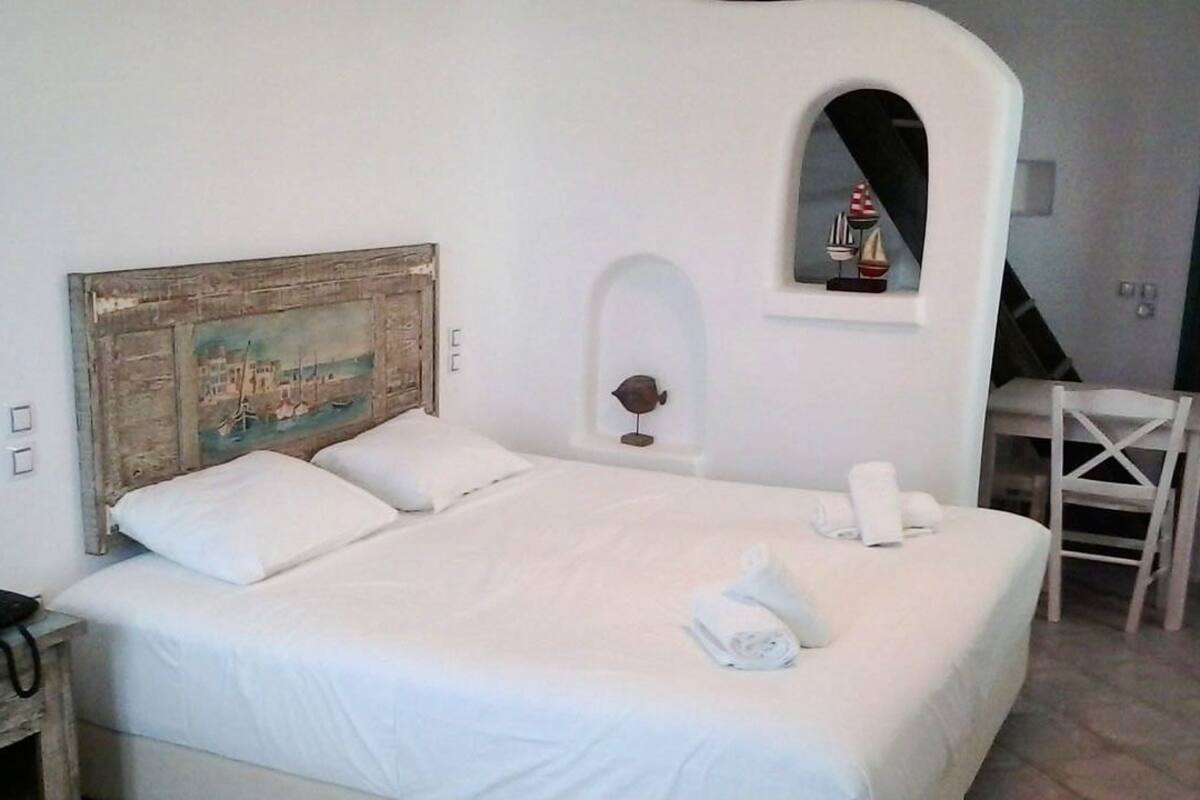 Ein Bett mit weißer Bettwäsche, ein Holzkopfbrett, ein kleiner Tisch mit Stuhl und ein an der Wand befestigtes Regal mit einer Dekoration.