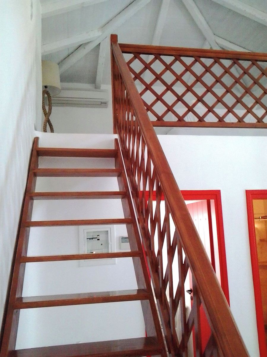 Escada de madeira com corrimão treliçado, paredes brancas e porta vermelha.