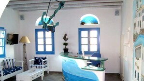 A room with a curved reception desk, two blue-framed windows, a white sofa with striped cushions, a small table, and a hanging light fixture. - Viva Mare Hotel (Pylos-Nestoras)