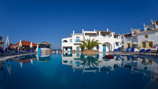 A clear blue swimming pool surrounded by white buildings with blue accents and palm trees. - Viva Mare Hotel (Pylos-Nestoras)