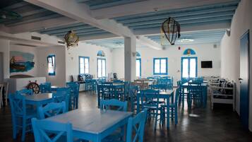 Een ruime eethoek met blauwe tafels en stoelen, witte muren en een hoge plafond met zichtbare balken.