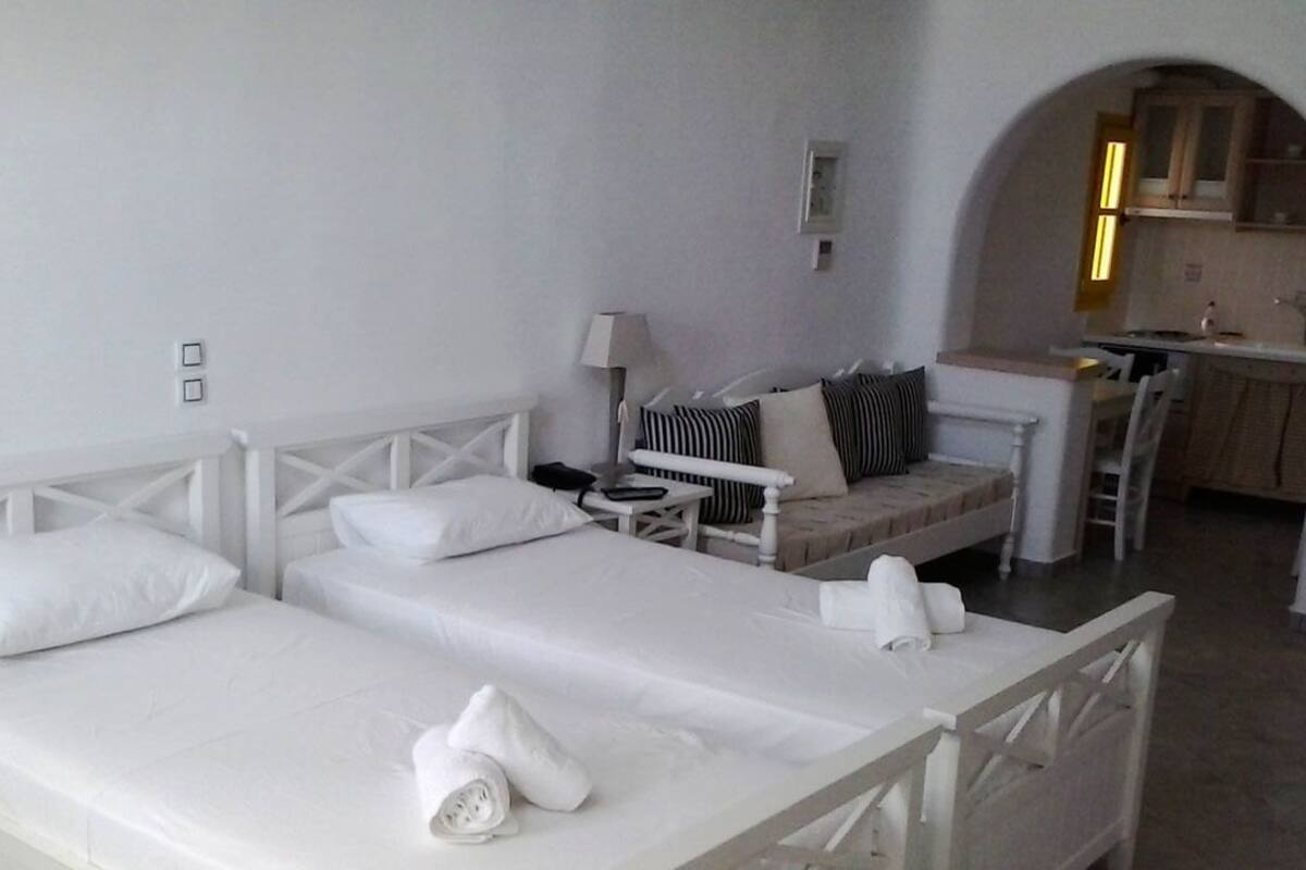 Ein Hotelzimmer mit zwei Betten, einem kleinen Tisch, einem Stuhl und einer durch einen Bogen sichtbaren Küchenzeile.