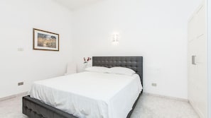 Un dormitorio con cama, mesita de noche, luz adosada a la pared y un cuadro enmarco.