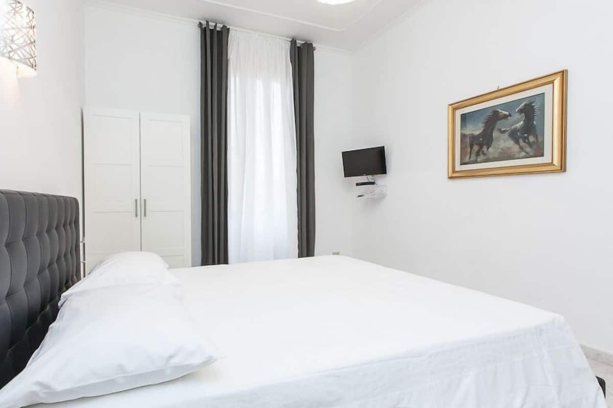 Une chambre à coucher avec une tête de lit capitonnée, un lit avec du linge de lit blanc, une armoire, un téléviseur fixé au mur et un tableau encadré.