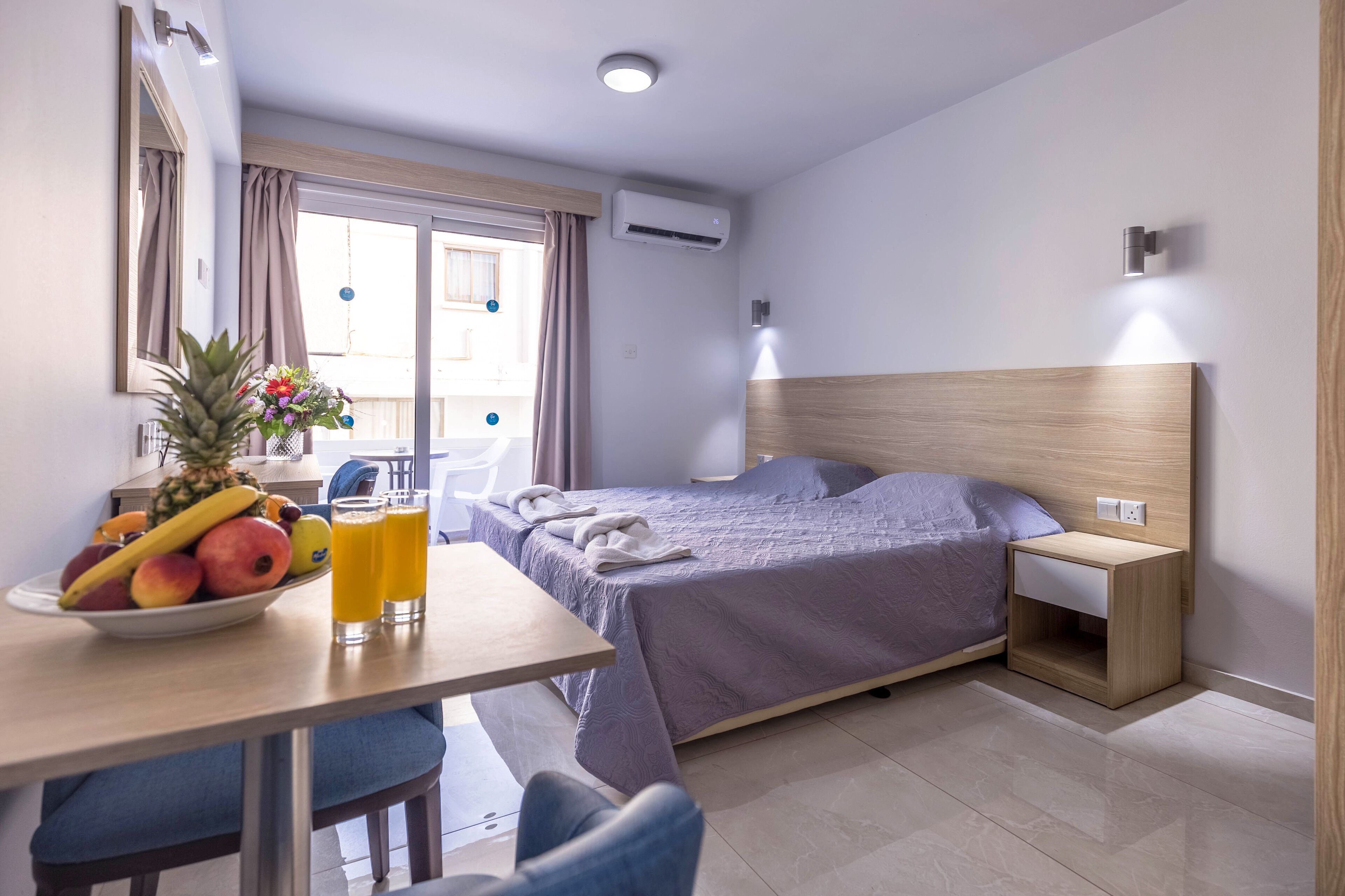 A hotel room with a bed, bedside table, and a dining area with fruit and drinks.