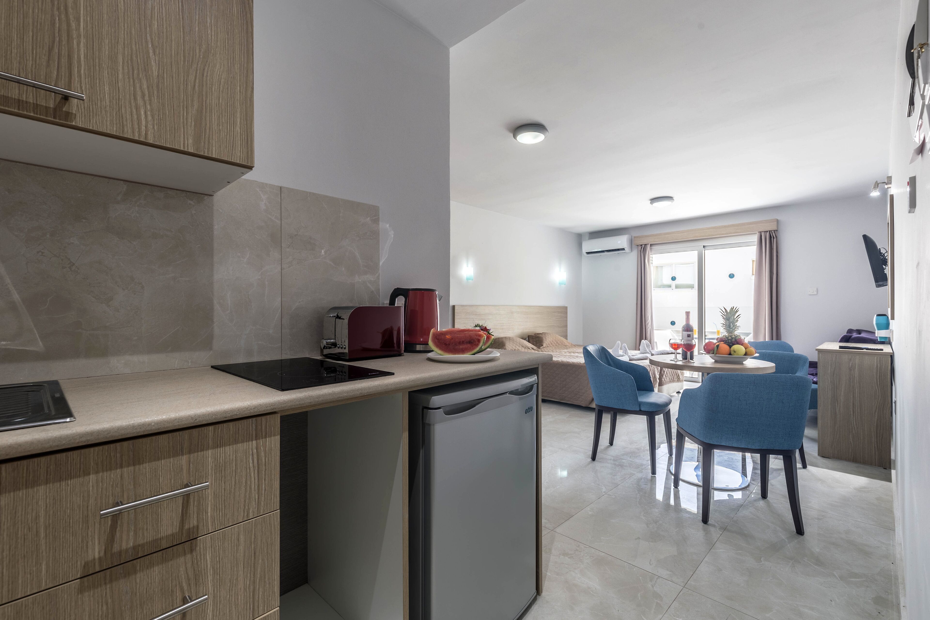 A modern kitchen with a dishwasher, a coffee maker, and a fruit bowl on the counter. There is a dining area with blue chairs and a table.