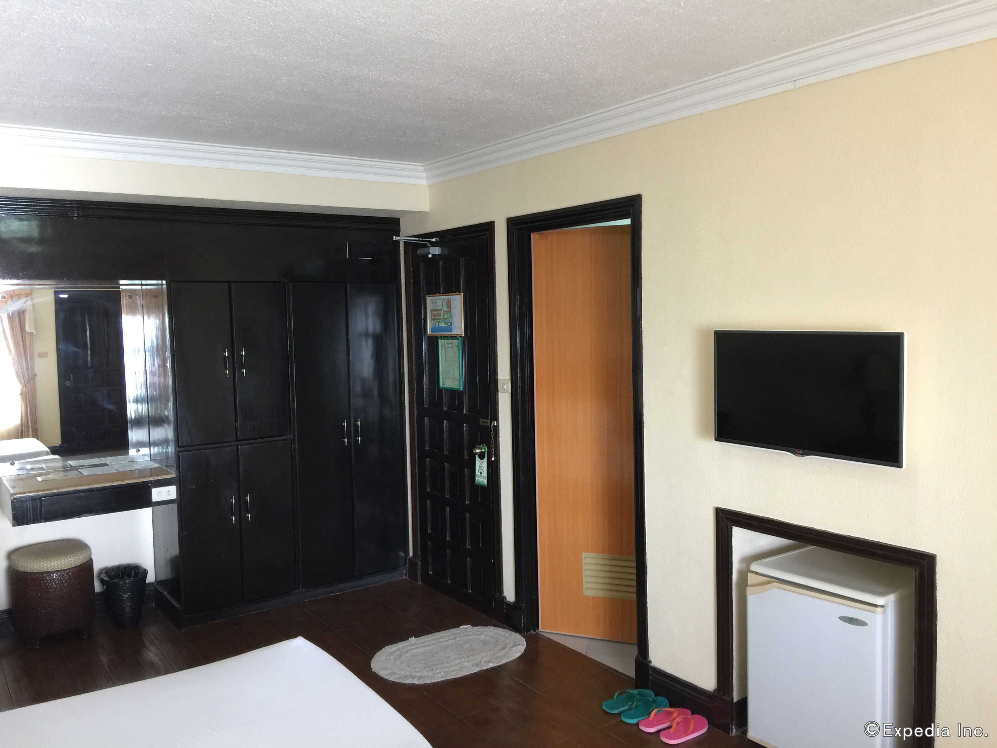 Une chambre d’hôtel avec un lit, un petit réfrigérateur blanc, un téléviseur fixé au mur et une porte en bois.