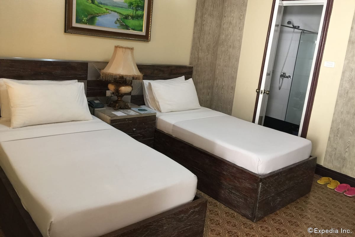 A hotel room with two single beds, a nightstand with a lamp, a painting of a landscape, and a shower area.